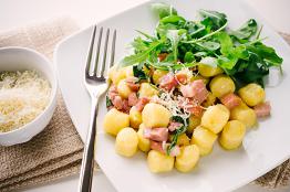 Gnocchi mit Speckwürfeln, Rucola und geriebenem Parmesan auf einem weißen Teller.
